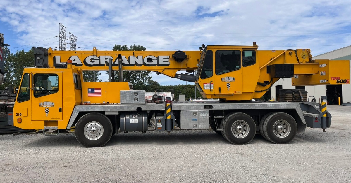 A yellow mobile crane parked on gravel beside a building, with trees, power lines, and a cloudy sky behind it.