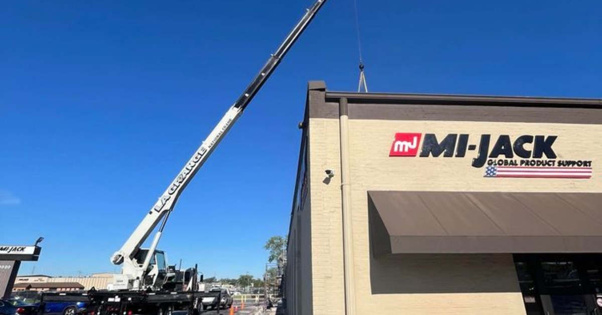 A large truck crane parked outside a commercial business during the daytime. The crane arm is raised in the air.