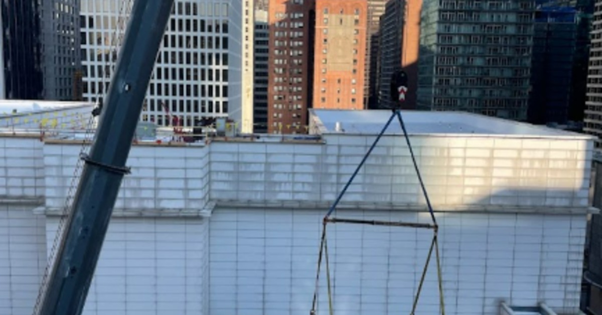 A large crane transporting gray metal machinery up the side of a tall skyscraper on a clear, sunny day.