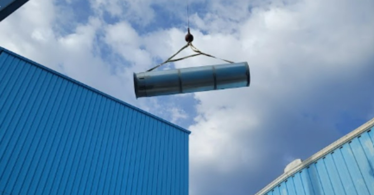 A crane carrying a long metal tube in the air above a blue metal building. The sky above the crane is cloudy.