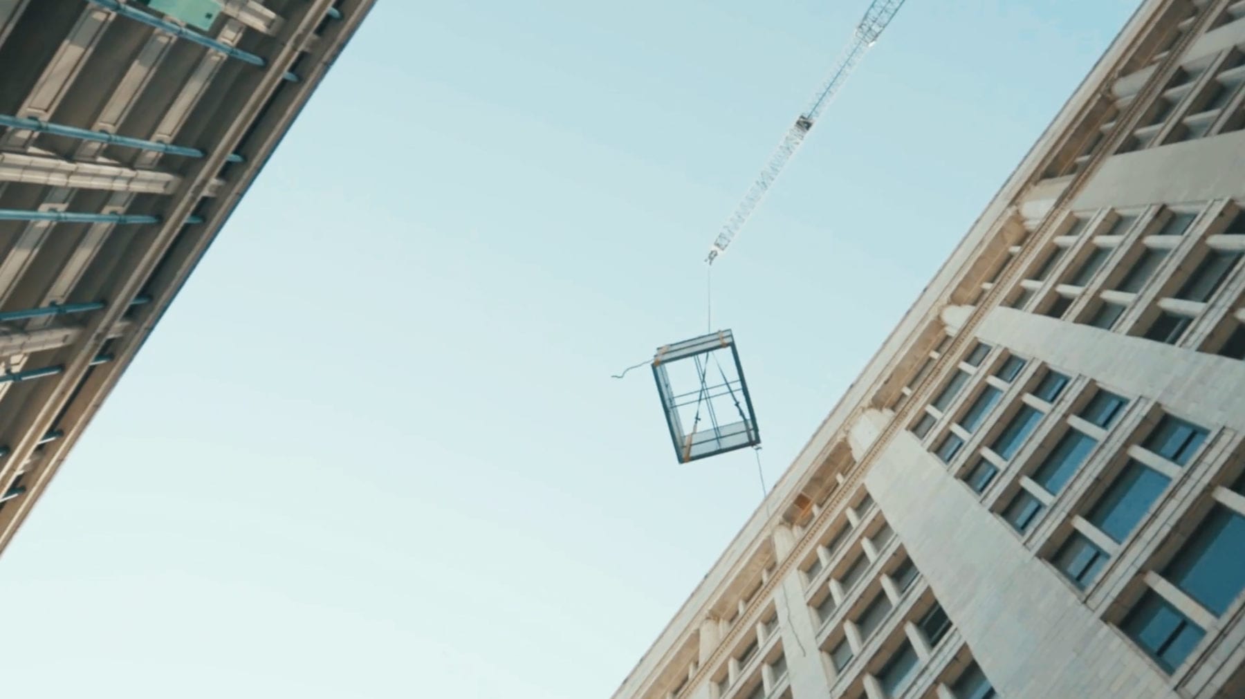 A yellow crane with its arm extended high into the air between two large buildings. A cage hangs from the boom arm.