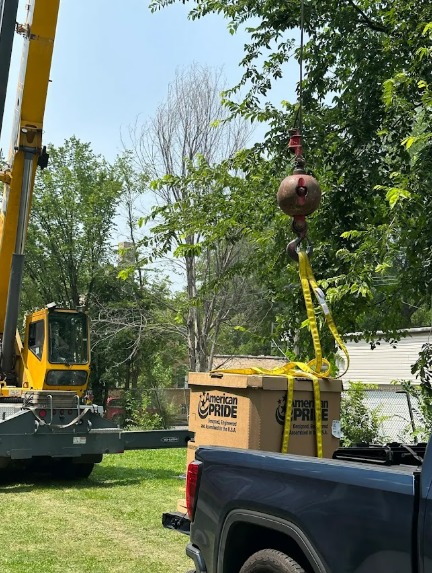 A yellow crane in a residential backyard during the daytime. A cardboard box hangs on the crane hook.