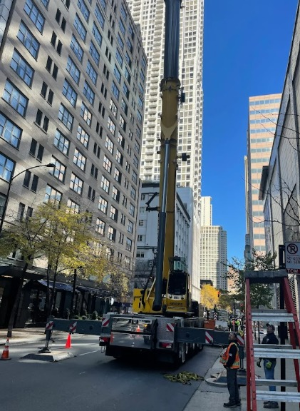 A tall yellow crane outside on a city street during the daytime. A ladder waits on the sidewalk nearby.
