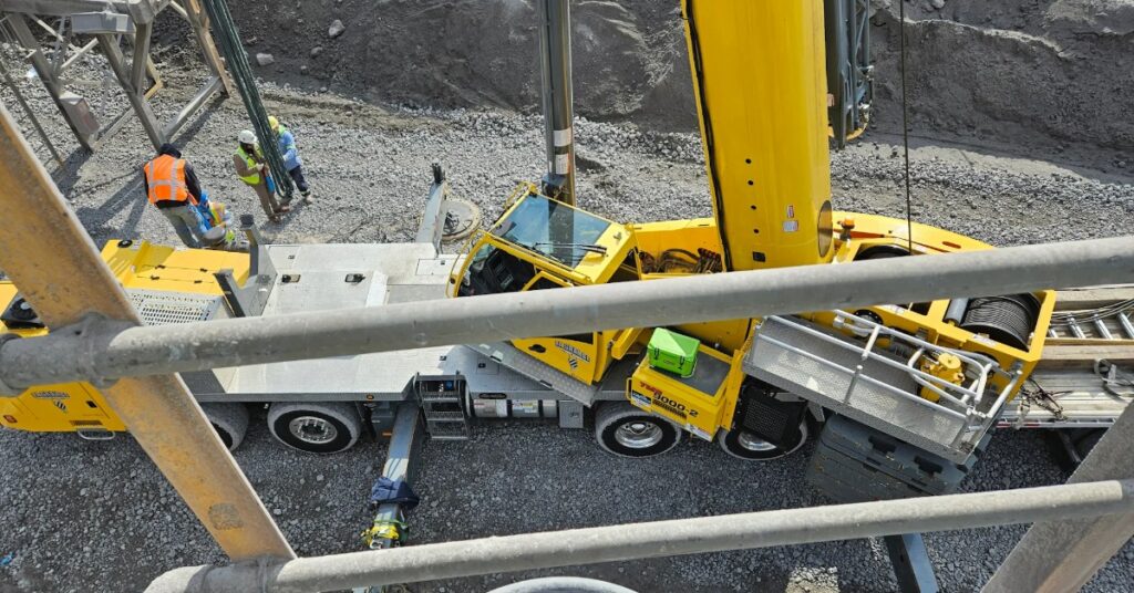 A yellow crane truck on a gravel worksite, viewed from above through railings with workers and equipment nearby.