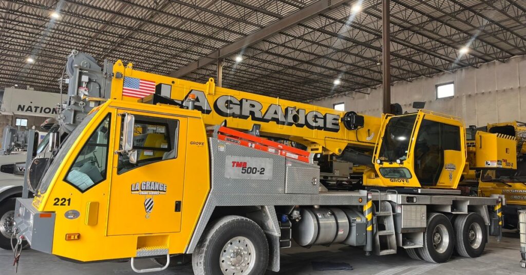 A long yellow all-terrain crane parked inside a large building with small windows. The crane is lowered.