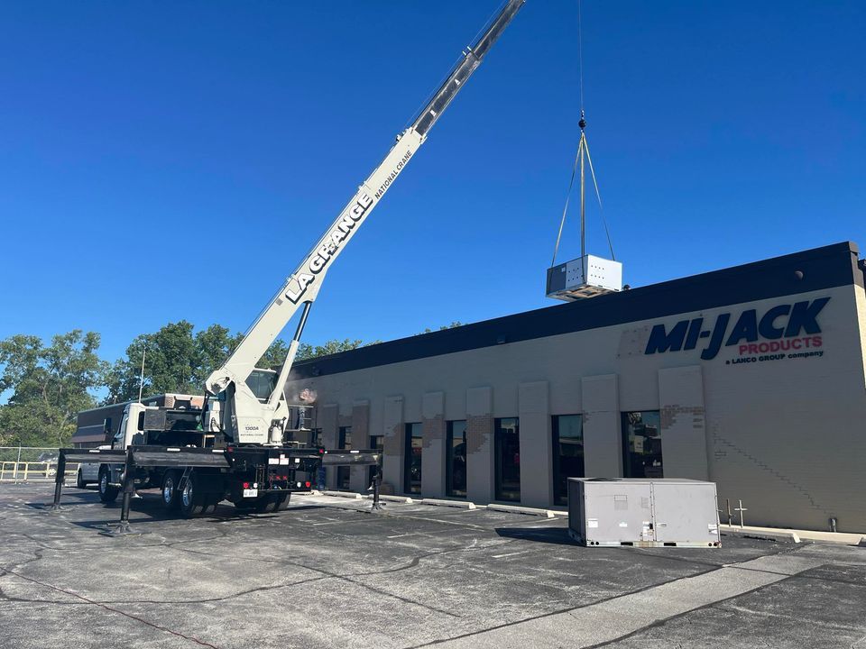 A large white truck crane lifting a small metal piece of machinery outside a store during the daytime.