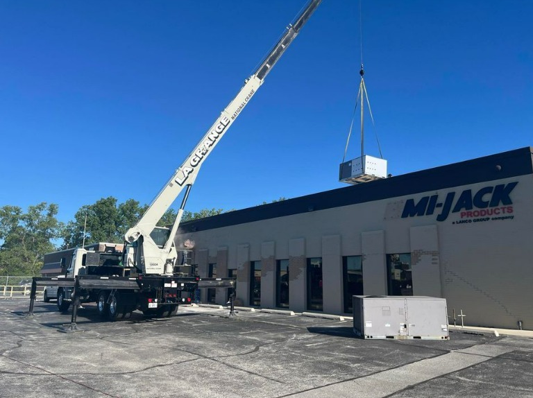 A large white crane carrying a bulky HVAC unit to the roof of a store. The crane reads "La Grange" on the side.